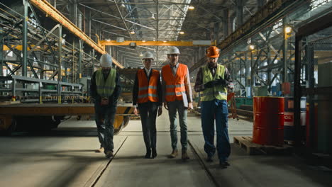 four heavy industry engineers discuss work process walking at manufacturing factory four heavy industry engineers in safety uniform and hard hats discuss work process walking at manufacturing factory mature supervisor holds laptop talks to diverse workers in warehouse slow motion