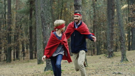 glücklicher kaukasischer vater, der versucht, seinen kleinen sohn zu kathodieren, während sie spielen, superhelden mit roten umhängen im wald zu sein