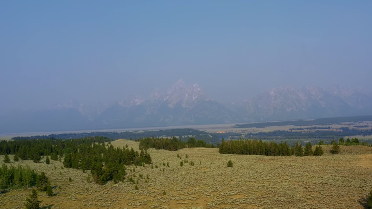 Aerial drone landscape nature tilting up dolly out shot of the majestic Grand Tetons National Park mountain range with a valley of brush and pine trees below on a warm hazy summer day in Wyoming, USA.