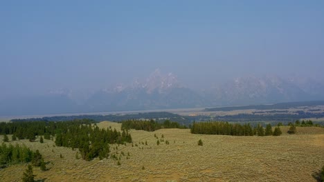 aerial drone landscape nature tilting up dolly out shot of the majestic grand tetons national park mountain range with a valley of brush and pine trees below on a warm hazy summer day in wyoming, usa.