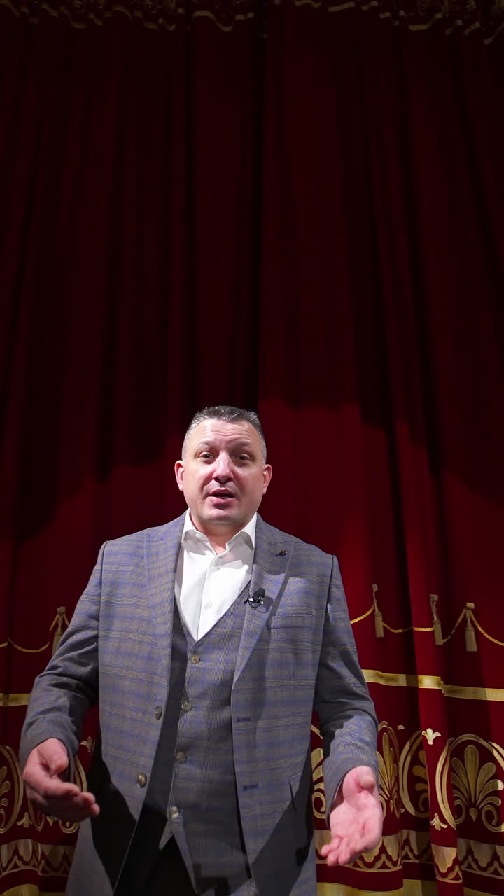 Man in gray costume on stage. Middle-aged actor talking on camera on luxury red curtains background in the theater. Slow motion. Vertical video