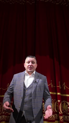 man in gray costume on stage. middle-aged actor talking on camera on luxury red curtains background in the theater. slow motion. vertical video