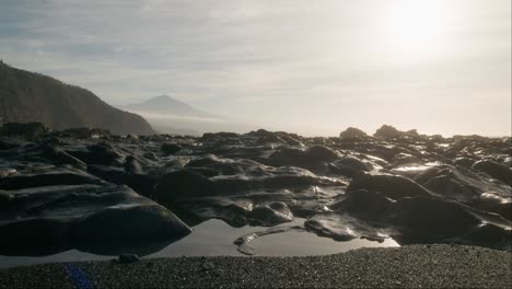 etérea playa de tenerife al amanecer con arena negra y rocas volcánicas y el brumoso pico del teide en el fondo, islas canarias