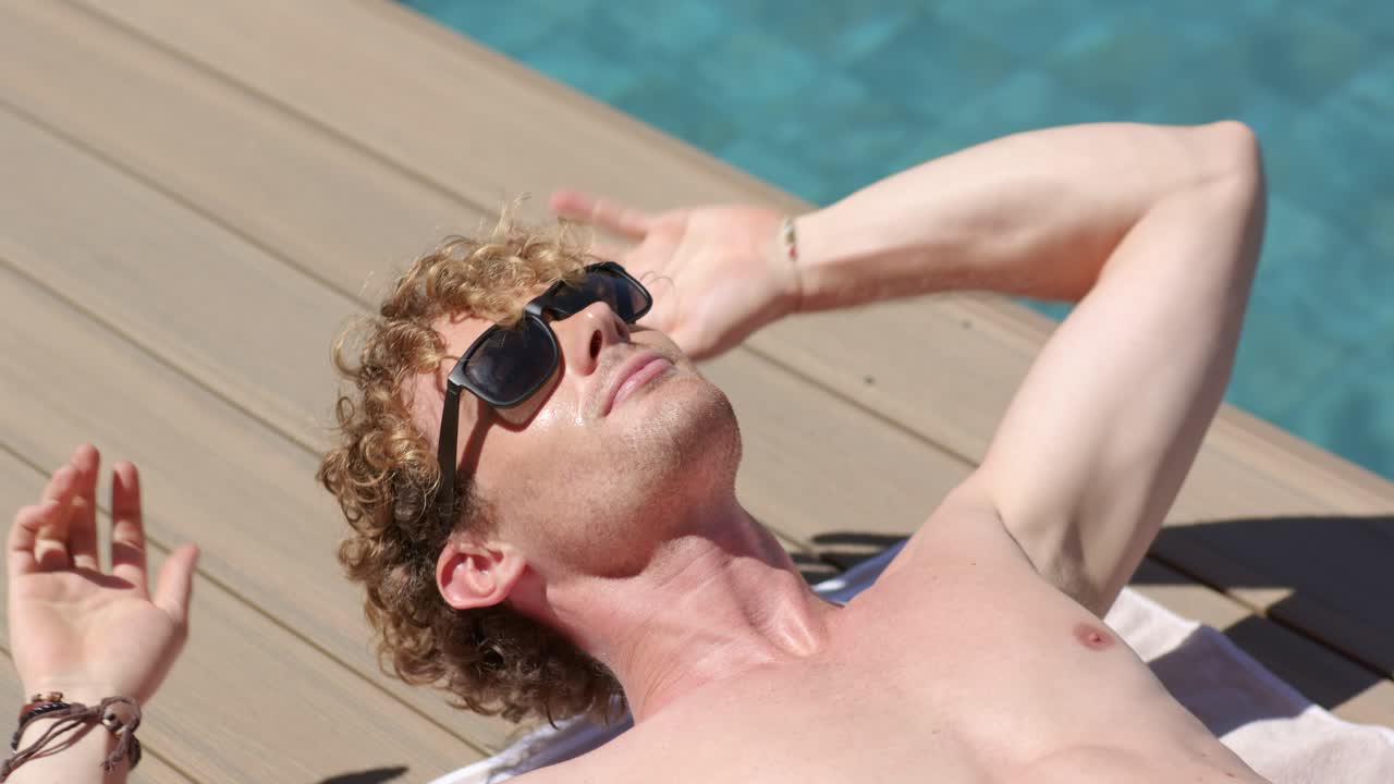 Uomo sdraiato su un asciugamano su una pedana di legno a bordo piscina, la telecamera arretra, lui si appoggia e indossa occhiali da sole.