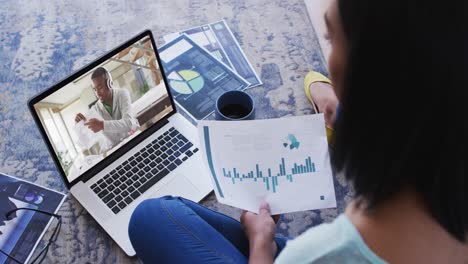 Mixed-race-businesswoman-sitting-on-floor-using-laptop-having-video-call-with-male-colleague