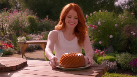 a smiling woman with red hair presents a bundt cake on a table in a garden