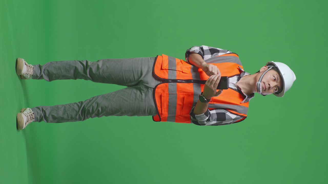 Full Body Of Asian Male Engineer With Safety Helmet Using Smartphone And Looking Around While Standing In The Green Screen Background Studio