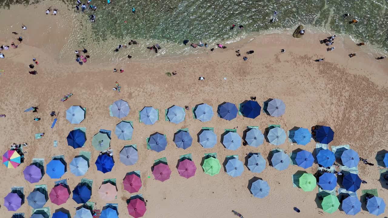 Aerial top-down view of colorful beach umbrellas arranged neatly on a sandy shore with people relaxing and swimming near the turquoise sea. Perfect summer vacation scene in tropical paradise