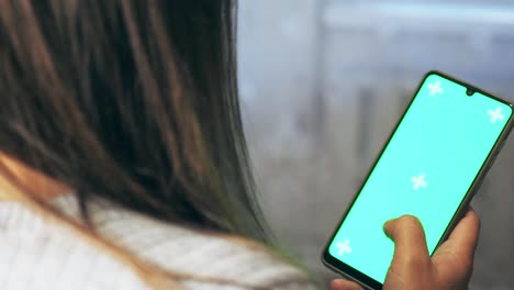 A girl with brown hair holds a phone in her hands with a green background. Back view. A woman's hand holds a mobile phone on the screen of which is a chroma key. A woman is scrolling on her phone.