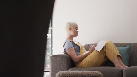 feliz mujer biracial tomando notas y sentada en el sofá en cámara lenta