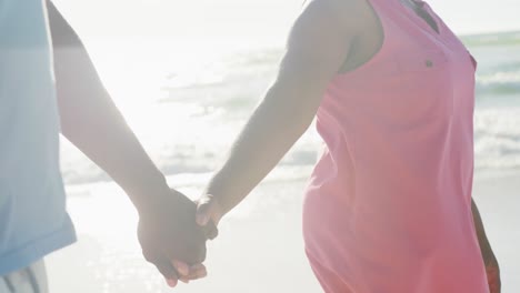 Midsection-of-senior-african-american-couple-walking-and-holding-hands-at-beach,-in-slow-motion