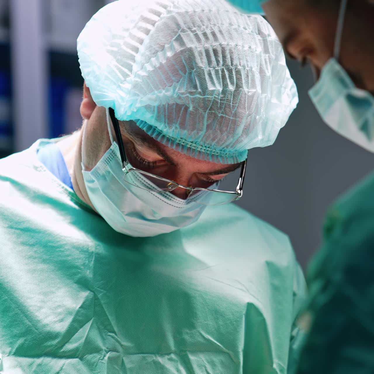 Un chirurgo di mezza età con cappellino, mascherina e occhiali guarda in basso mentre opera il paziente. Primo piano di un dottore al lavoro