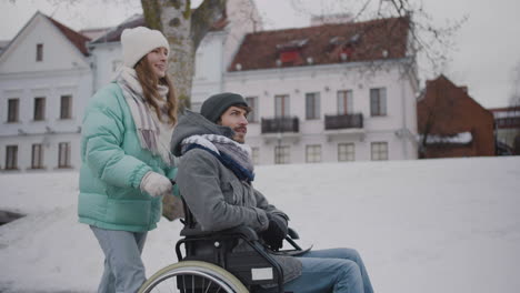 vista lateral de una mujer caucásica feliz llevando a su amiga discapacitada en silla de ruedas a dar un paseo por la ciudad invernal y hablando juntos