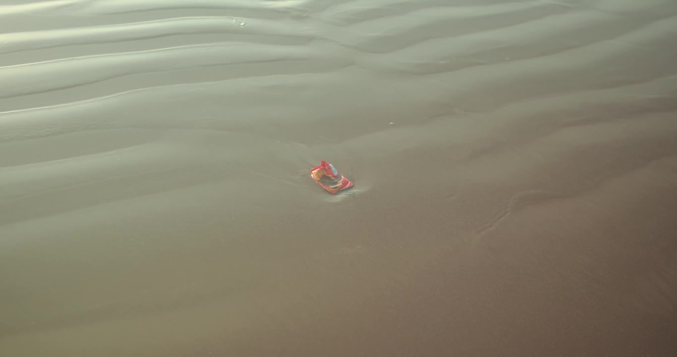 Small red object resting on rippled wet sand at low tide, surrounded by soft textures and minimal patterns. Abstract coastal detail suited for backgrounds, environment and conceptual visuals.