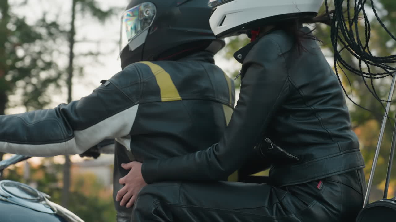 Back view of motorcycle rider and passenger wearing helmets and black leather gear traveling on road, long braids of passenger visible, with nearby car and trees in soft evening light background