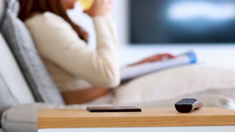 an asian woman sits on a modern sofa, drinking juice and watching television in a bright, cozy living room with soft natural lighting