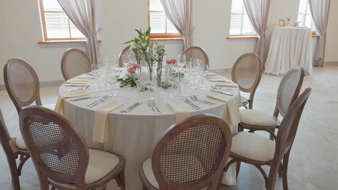 Bright, airy event space with round tables dressed in white linens, elegant settings, fresh flowers, and soft natural light. Wide-angle pan reveals tastefully decorated venue, no people, modern minima