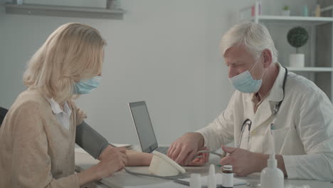 a grey haired, middle aged doctor in a facemask doctor takes blood pressure to a female patient