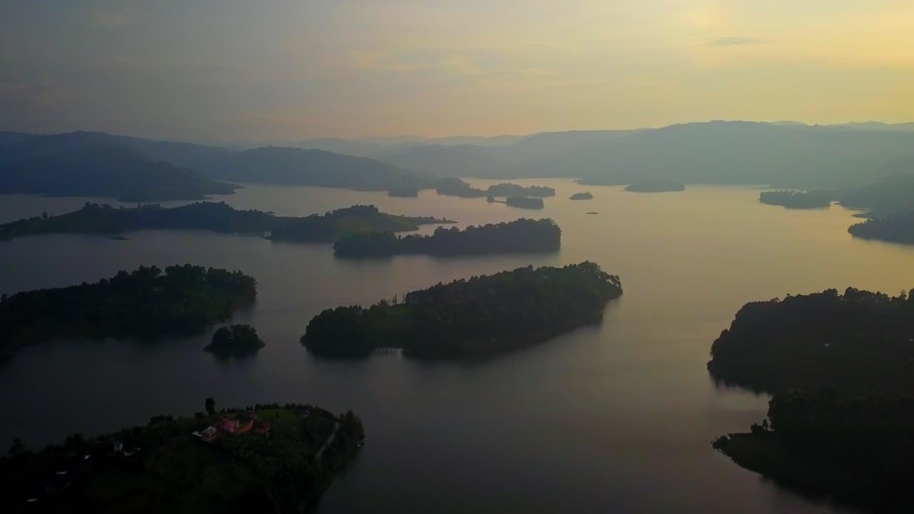 Aerial drone view of a golden sunset casting reflections across the calm waters of Lake Bunyonyi, Uganda, with silhouettes of multiple small islands and rolling highland terrain in the distance