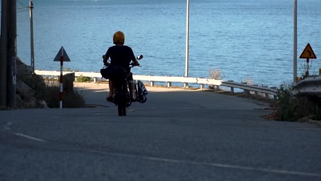 Man-rides-motorbike-with-surfboard-on-rack-on-seaside-road,-kiteboarding-trip