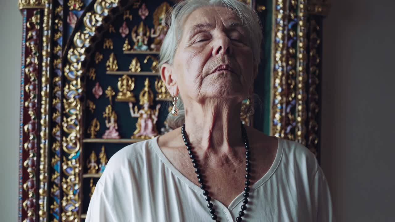 Elderly woman with serene expression engages in meditation practice, surrounded by intricate decorative backdrop, embodying tranquility and mindfulness in a spiritual setting