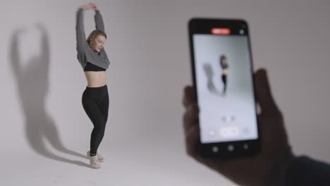 person recording video on mobile phone of woman doing dance practise on grey background in studio