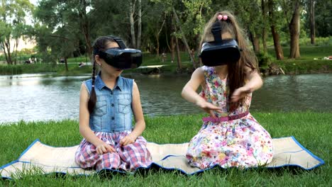 two little girls play virtual games in the park on the river bank. they sit in virtual reality glasses, look around and wave their hands.