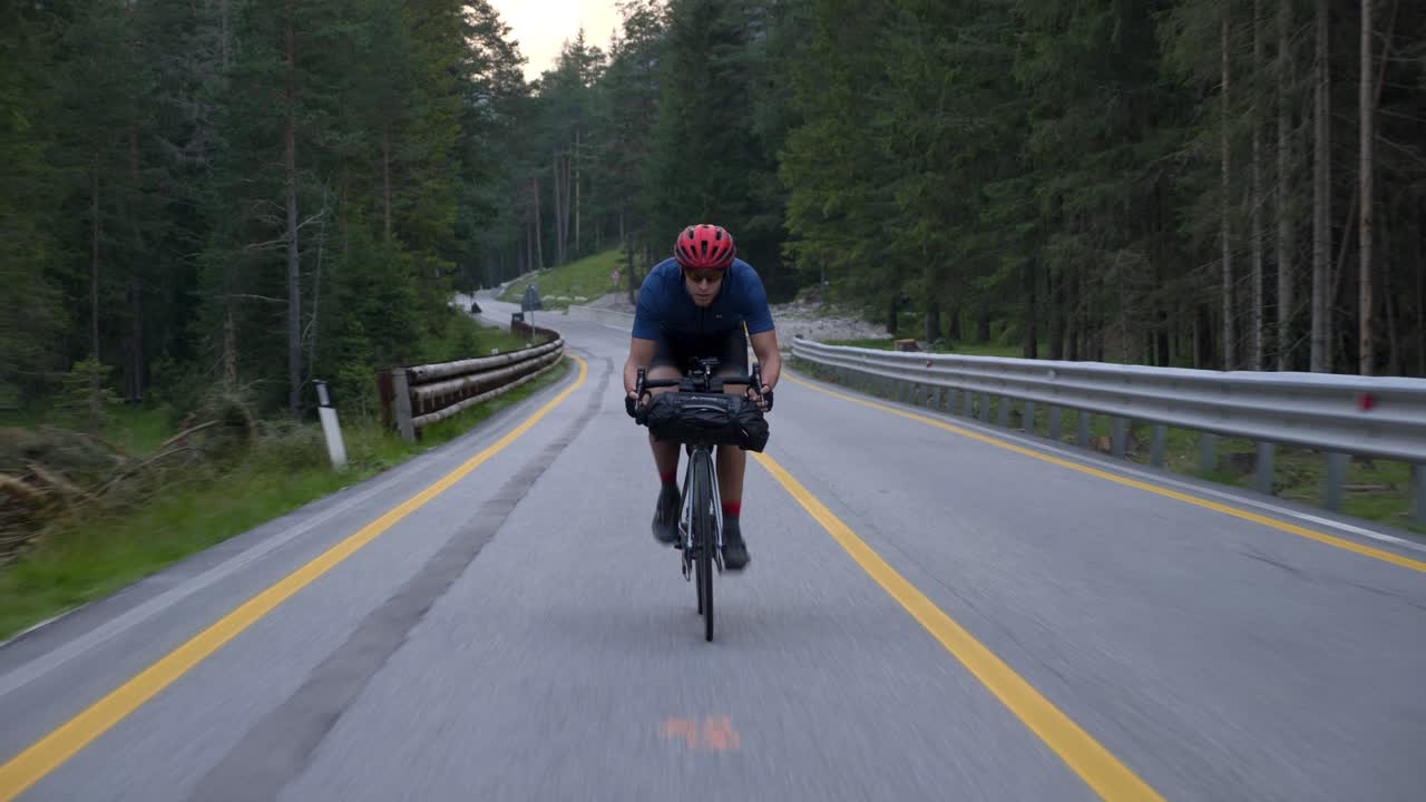 Ciclista con casco rosso che pedala su strada asfaltata tra le montagne con linee gialle in una pineta, Dolomiti, Italia.