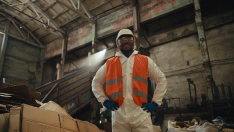 A-confident-and-happy-man-with-Black-skin-in-a-white-helmet-and-a-special-protective-uniform-stands-near-a-large-pile-of-garbage-and-a-conveyor-at-a-huge-waste-recycling-plant