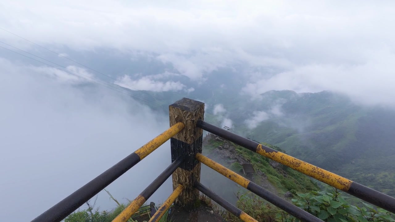 A breathtaking cinematic reveal shot of showing thick clouds rolling over the lush green hills of Meghalaya, captured from a stone viewpoint with safety railings.