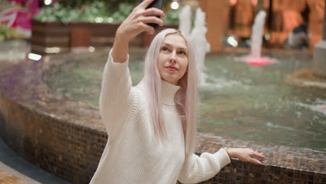 stylish selfie seeker sits beside ornate mall fountain, manicured hand skims water surface while she raises phone to frame smiling portrait, blurred children play near neon lit shops in concourse