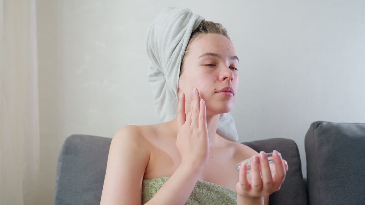 Young woman wrapped in towel with hair covered in headwrap applies skincare cream to face using fingers while sitting on couch near window in soft daylight during peaceful home beauty routine