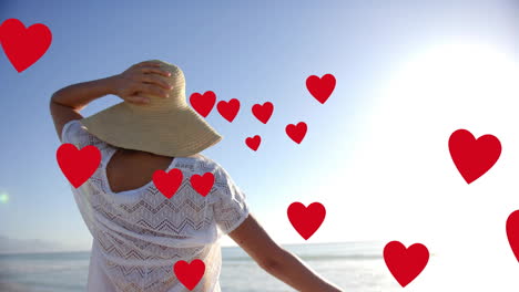 animation of hearts moving over biracial woman in hat on beach in summer
