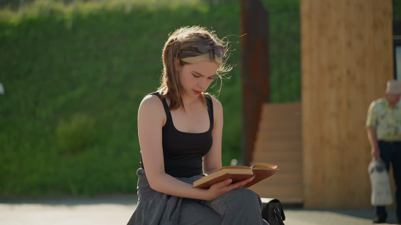 Lady seated with legs crossed, reading a book while wind blows her hair, her hand rests on the book, a black bag is nearby, with a blurred background featuring a man and woman in the distance