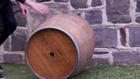 rolling a wine wooden barrel across green grass past a stone wall