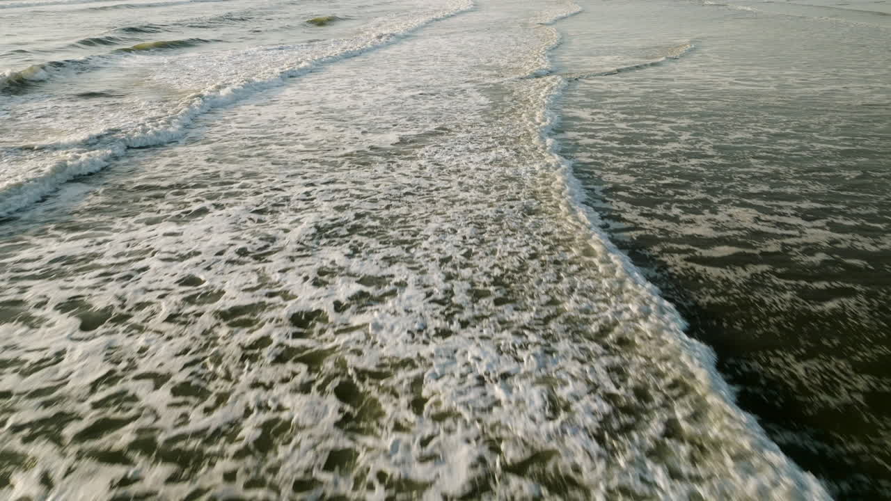 Aerial dolly perpendicular to open ocean waves crashing as whitewash spreads rolling to beach, natural background