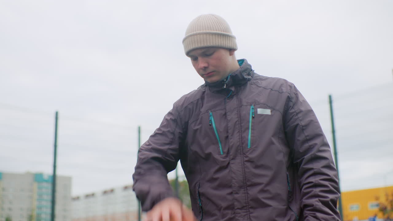 Young man dribbling basketball outdoors on overcast urban court, wearing insulated jacket and knit beanie, metal fence and apartment blocks in background, slow focused practice showing calm