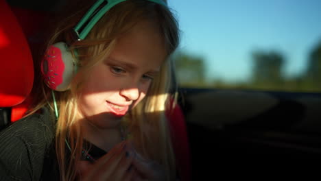 blonde schoolgirl wearing headphones sitting relaxed in backseat, gazing down while listening to music during family road trip across scenic countryside landscape