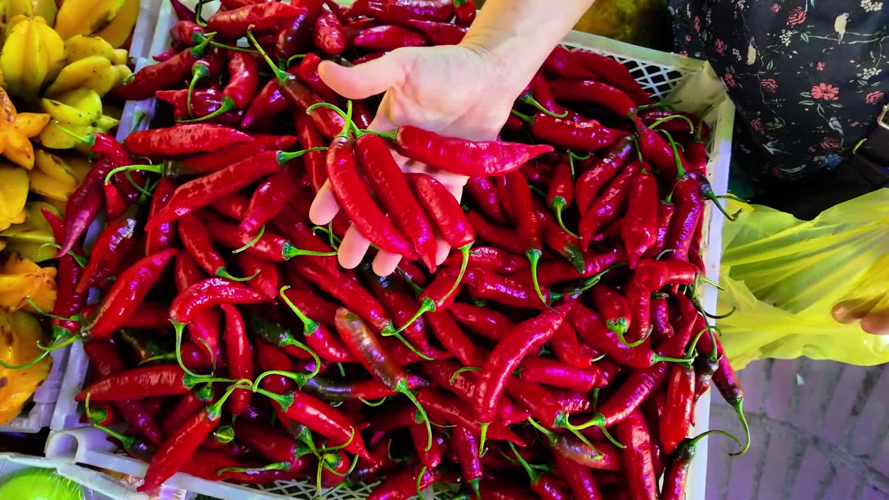 Hands are selecting bright red chili peppers from a basket in a lively market. The atmosphere is vibrant with fresh produce, showcasing the rich colors and textures of the ingredients available