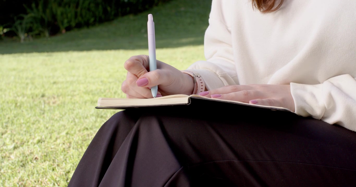 Pen poised, woman is journaling on lap in park with left hand holding notebook page