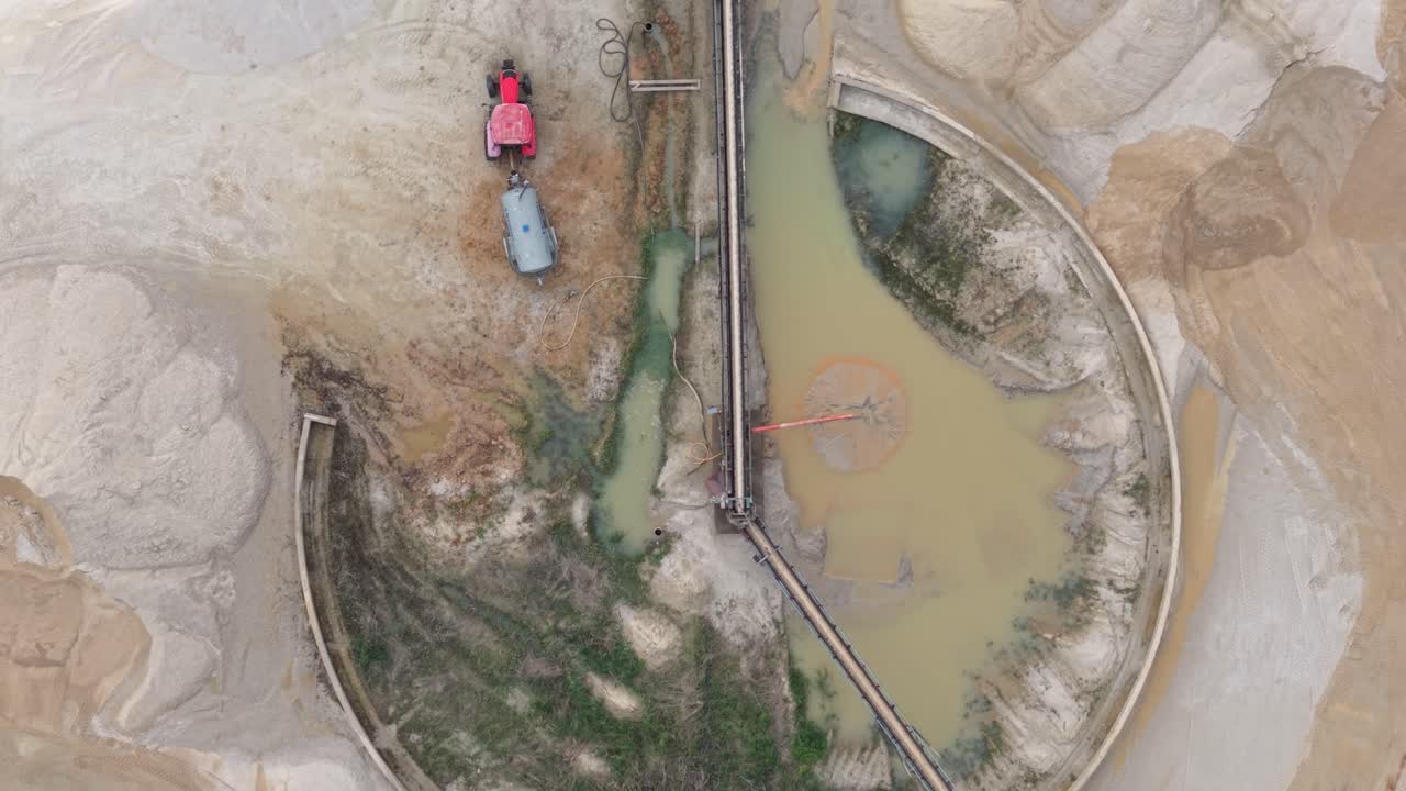 A top-down view of a large circular sedimentation basin filled with murky water and connected to conveyor belts. Surrounding the basin are sand piles and traces of vegetation,