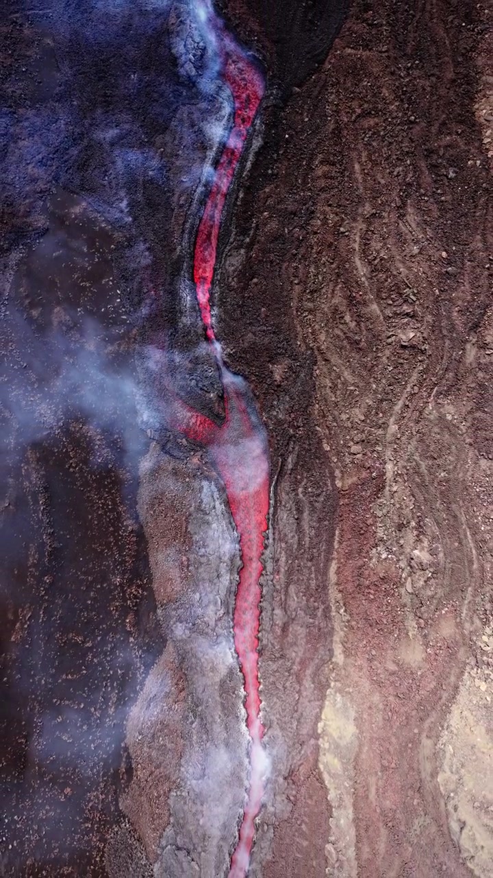 Pandangan udara vertikal sungai lava pijar mengalir di antara batuan vulkanik gelap dan uap