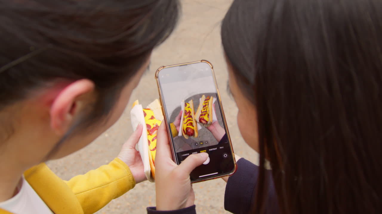dos amigas tomando fotos de perritos calientes comprados en un puesto de comida callejera en un teléfono móvil 1