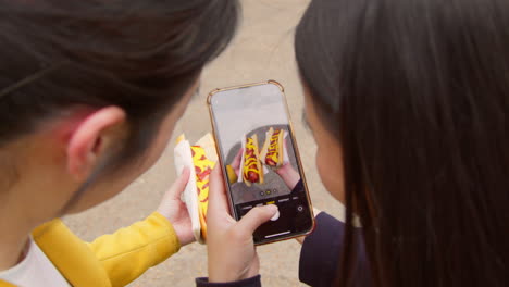 Zwei-Freundinnen-Fotografieren-Mit-Dem-Handy-Hotdogs,-Die-Sie-An-Einem-Straßenimbissstand-Gekauft-Haben-1