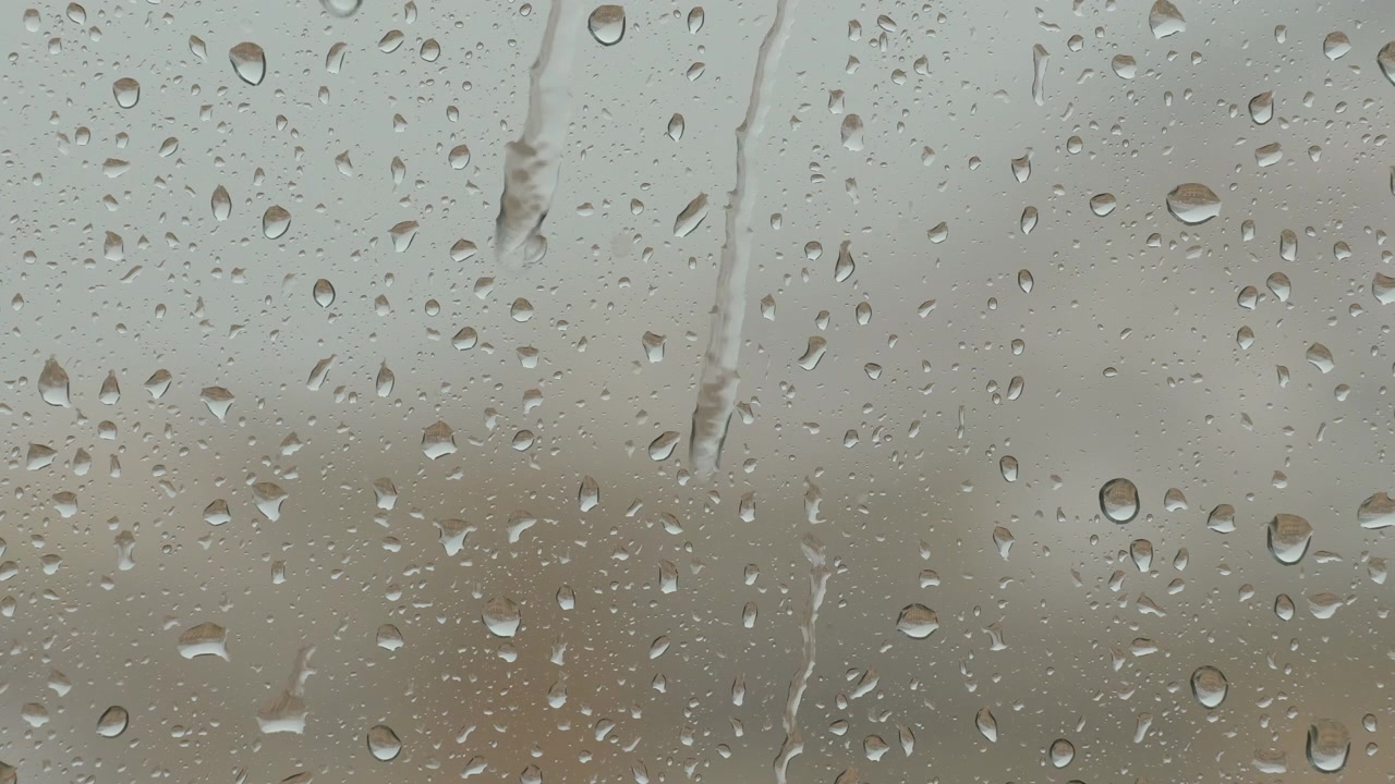 Primo piano di goccioline di pioggia su un vetro di finestra con uno sfondo sfocato. Luce naturale diffusa che crea un'atmosfera malinconica, nostalgica e calma. Elevato dettaglio della texture e del movimento dell'acqua.