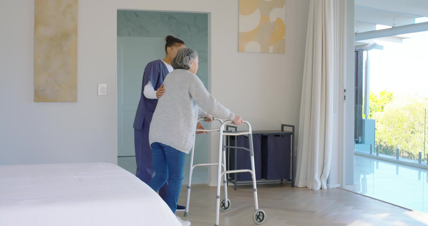 Pleier går inn på soverommet, støtter eldre person som bruker rullator mot skyvedør i glass for å se ut