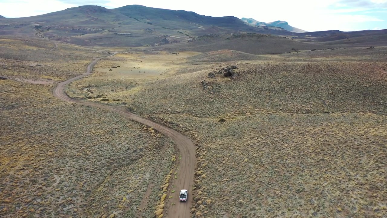 Ripresa con drone dall'alto di un veicolo 4x4 bianco che percorre un sentiero sterrato tortuoso attraverso una vasta valle di montagna a Bariloche, Argentina. Evidenzia viaggi remoti, esplorazione avventurosa e natura selvaggia.
