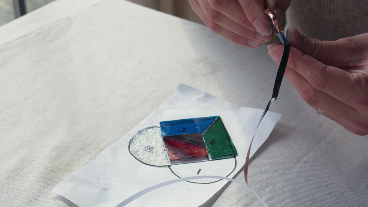 Um estudante de arte masculino descola fita de cobre sobre um design de vitral durante uma pequena aula de arte, sentado junto a uma janela