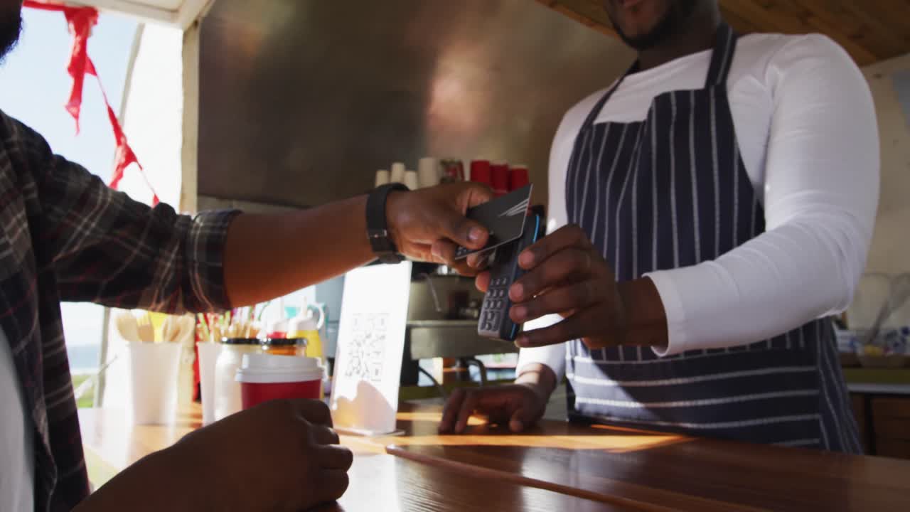 Afroamerikansk mand betaler den mandlige ejer for kaffe med kreditkort ved food trucken.