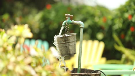 water from a garden spout pours and spills over a hanging bucket into a larger barrel with colored lounge chairs in the background